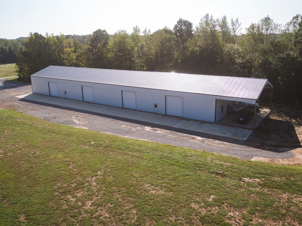 Aerial view of a large, rectangular white warehouse with a metal roof, surrounded by trees. The building has several closed doors and a covered area with a parked vehicle. The foreground has grass, while the background features leafy trees under a clear sky.
