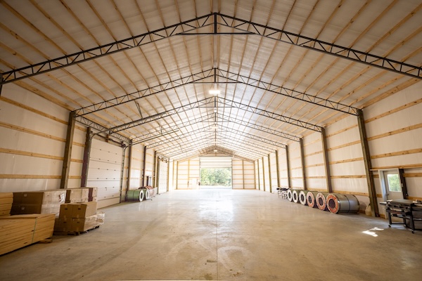 Interior of an empty warehouse featuring metal support beams, a concrete floor, and stacked materials on the sides.